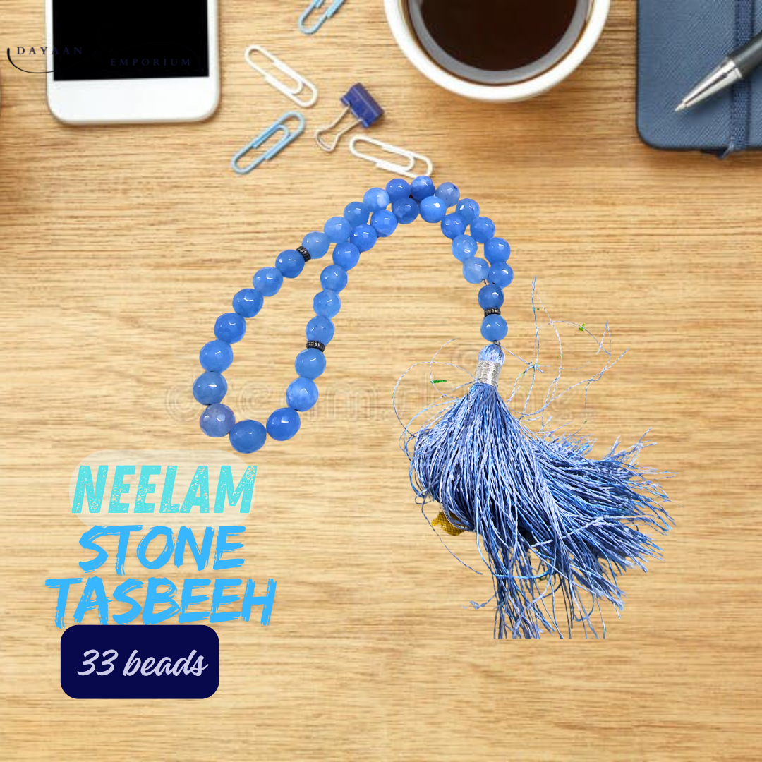 Blue tasbeeh with tassel on a wooden surface with smartphone, coffee, and stationery items.